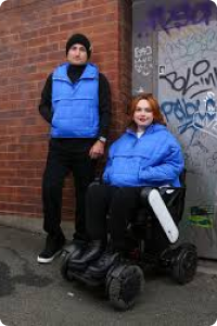 Man is standing wearing bright blue gillete and black hat and woman with red hair in a wheelchair wears jacket in same bright blue